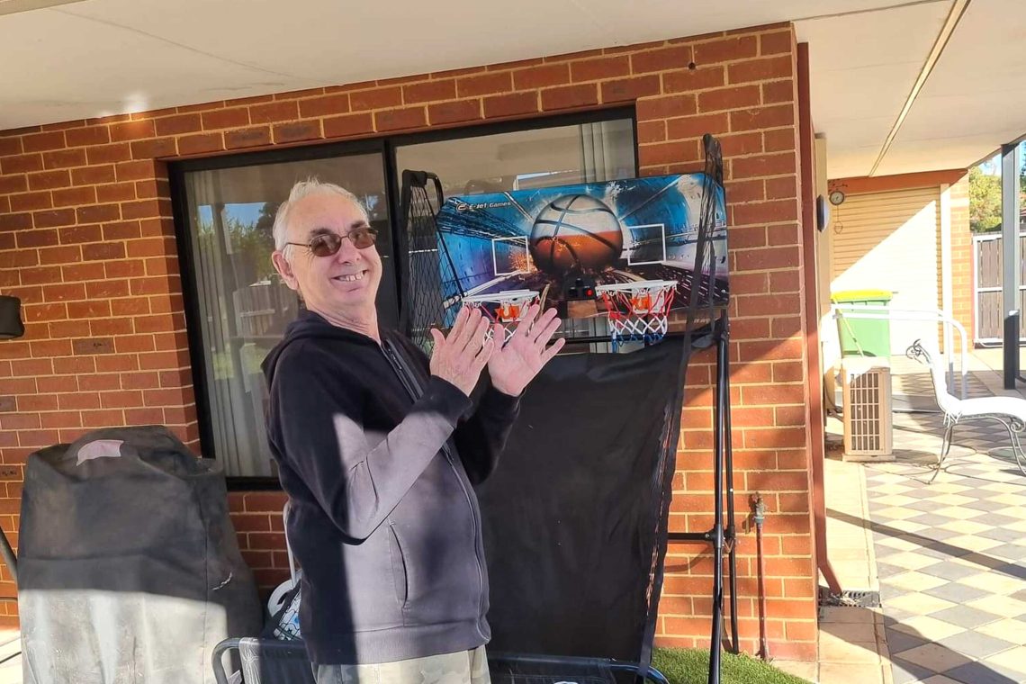 Older male smiling, standing in front of a small basketball hoop structure.