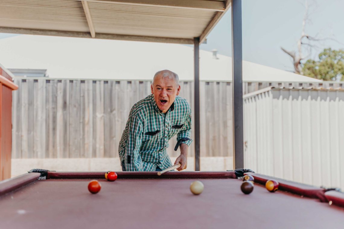 Older male playing billiards. He is leaning over slightly, with pool cue in one hand and a happily surprised expression on his face.