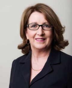Julie Waylen wearing a black blazer, gold loop earrings and squarish glasses with black frames, looking at the camera and smiling
